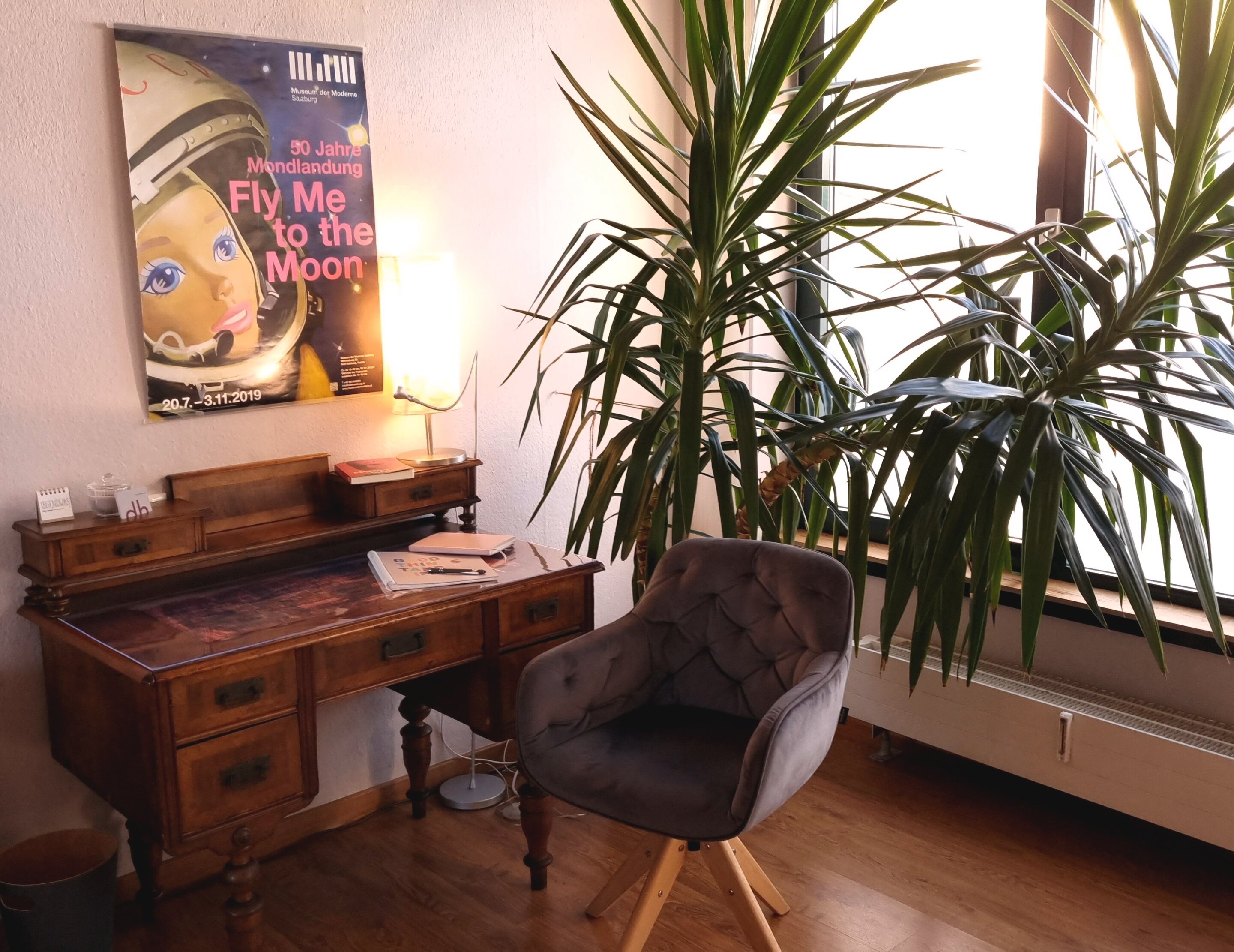 Ein alter Holzschreibtisch, davor ein grauer, gepolsteter Sessel. Im Hintergrund scheint das Sonnenlicht beim Fenster durch eine Palme herein.